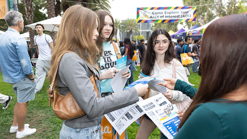 Students gathered around one student with long brown hair, facing the three female students reading campus maps. They are outside at a busy event.