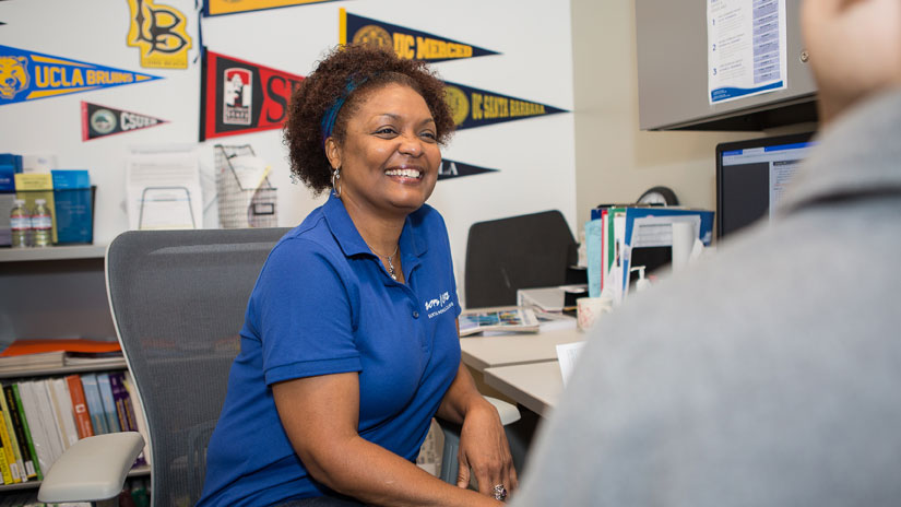 A female smiling councelor is sitting at her desk look and smiling at a student that is facing her. College pendents are hanging behind her.