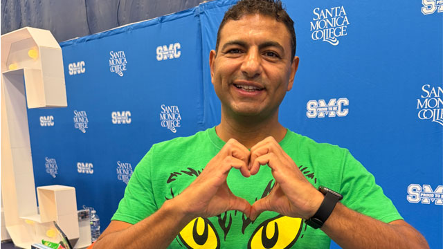 A man smiling at the camera, gesturing a heart with his hands. He wears bright, great t-shirts with printed black eyebrows and yellow eyes