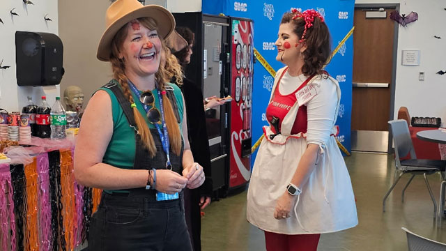 Two woman in the forground of the staff lounge the first woman laughing where scarecrow costume, the woman behind is wearing a red and white raggedy doll costume. 