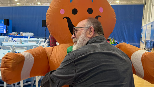 A man from behind, looking to the left, wearing glasses and has a grey beard, is hugging an inflatable gingerbread man 